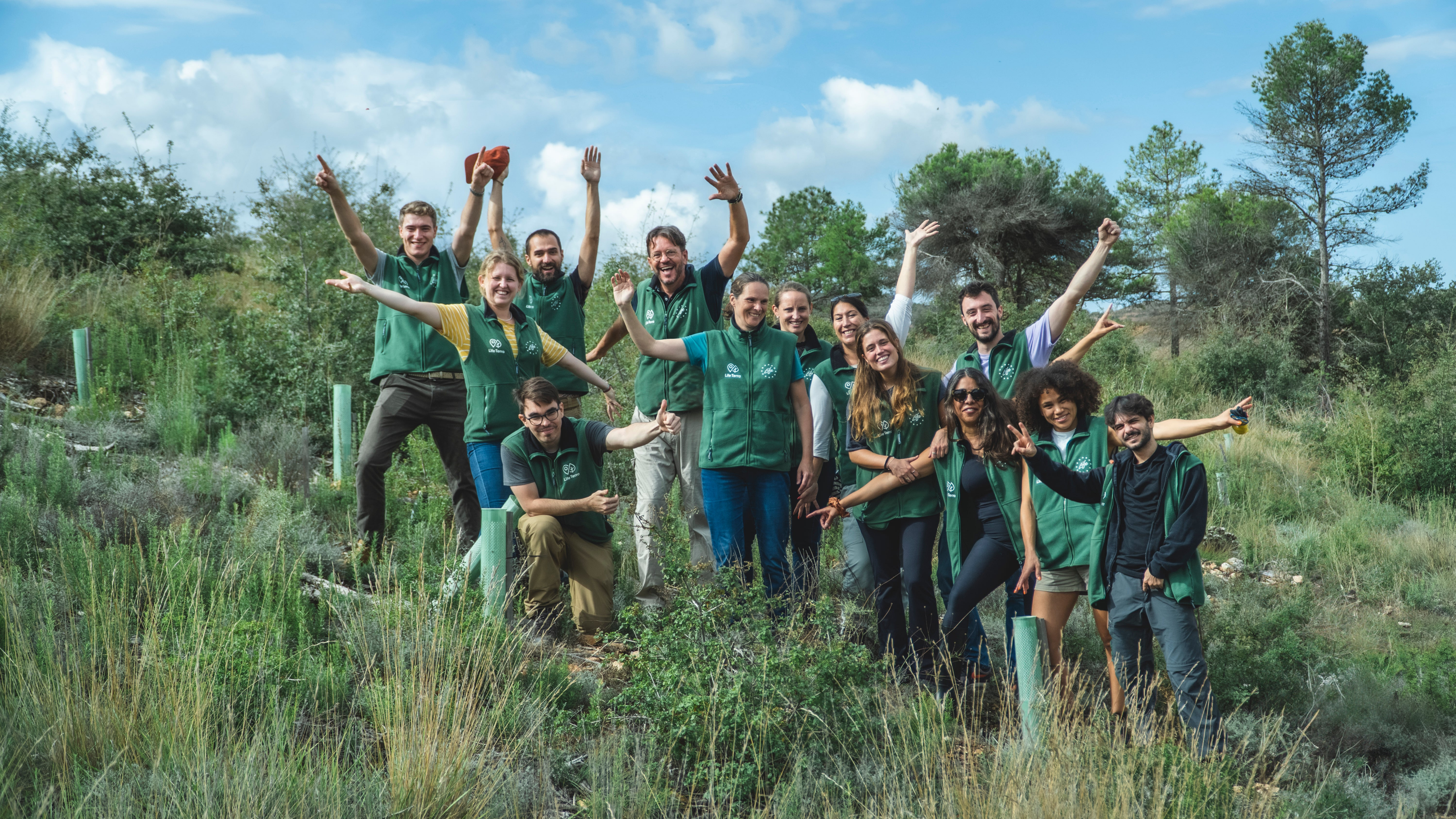 The Team of Life Terra, about 1 dozen people of mixed nationalities, standing on a green hillside with freshly planted trees, cheering with their hands in the air.